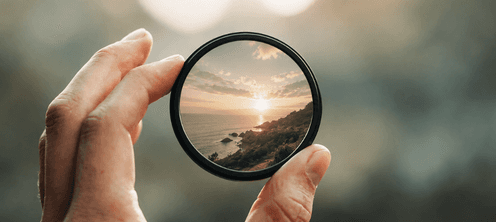 A hand holding a magnifying glass that brings a coastal sunset into focus