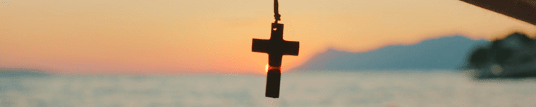 A cross pendant silhouetted against a sunset over the sea