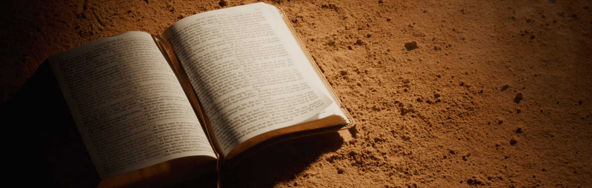 An open Bible resting on sandy ground, lit by warm sunlight